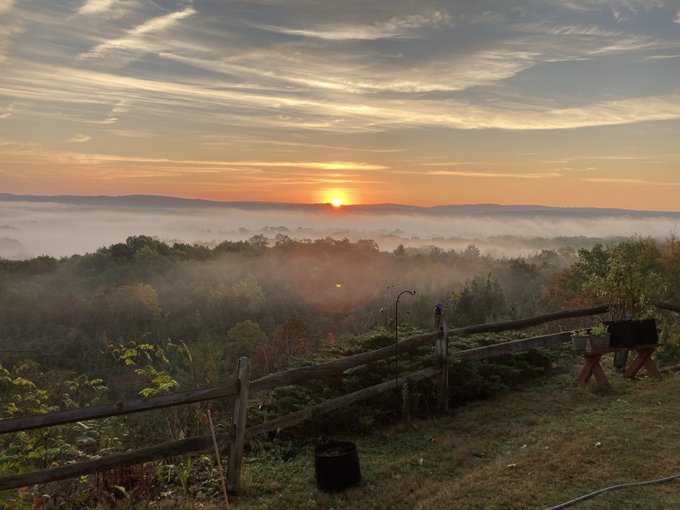 A scenic sunrise over a misty, forested valley, viewed from behind a rustic wooden fence and a small table with chairs.