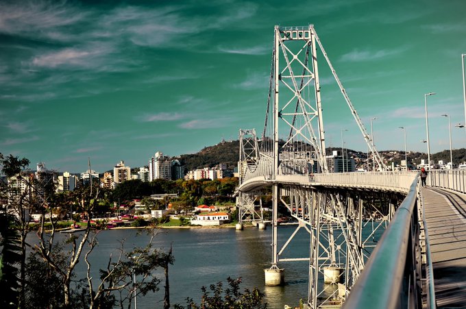 A dramatic, stylized photograph captures the Hercílio Luz Bridge in Florianópolis, Brazil, its towering steel suspension structure dominating the scene against a vivid teal sky and calm bay waters, with the city's hillside skyline stretching behind it. The image's artistic color grading and composition embody the post's message that "Photography is art," transforming a familiar landmark into a striking visual piece shared on the objkt.com NFT marketplace.