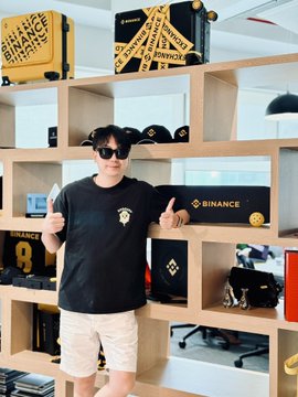 A person wearing sunglasses and a black t-shirt stands in front of shelves displaying Binance branded merchandise and boxes.