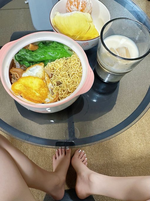 An overhead view of a meal on a glass table featuring a bowl of ramen with fried eggs and greens, a bowl of mango, a glass of milk with ice, and the diner's bare feet visible at the bottom.