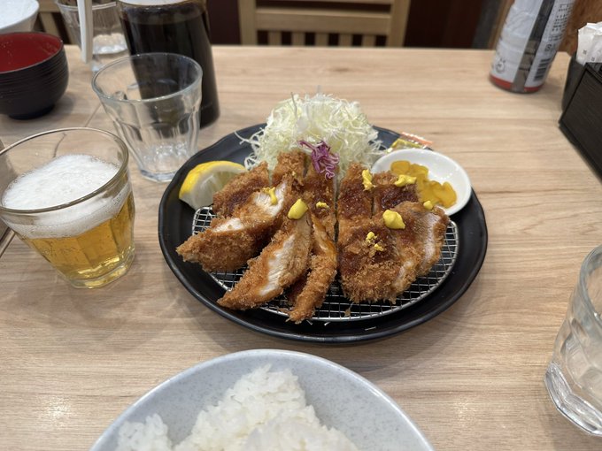 A Japanese meal featuring tonkatsu (breaded cutlets) topped with yellow mustard on a wire rack, served with shredded cabbage, rice, dipping sauce, and a glass of beer.