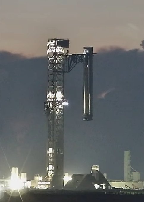 A SpaceX Starship V3 Super Heavy booster stands vertically on the launch pad at sunset, illuminated by bright floodlights, following its successful first-attempt catch. The towering rocket's stainless steel exterior gleams against the dusky sky, marking a major milestone in reusable launch technology.