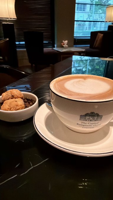 A frothy cappuccino served in a white cup branded "The Capitol Kempinski Hotel Singapore" rests on a saucer beside a small dish of chocolate chip cookies and other treats, suggesting a relaxing morning "GM from Singapore."