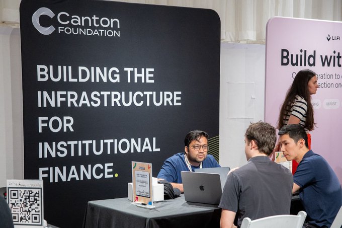 The image shows a group of people sitting around a table, engaged in conversation while using laptops. The table has a banner that says "Building the Infrastructure for Institutional Finance" written on it. There are four individuals visible in the scene - two men and two women. One man is seated at the head of the table, while another man sits next to him. A woman is also present near them.

The laptops they're using suggest that they might be discussing or working on financial applications related to privacy-first finance. The image captures a moment where these individuals are collaborating and sharing ideas in an indoor setting.
