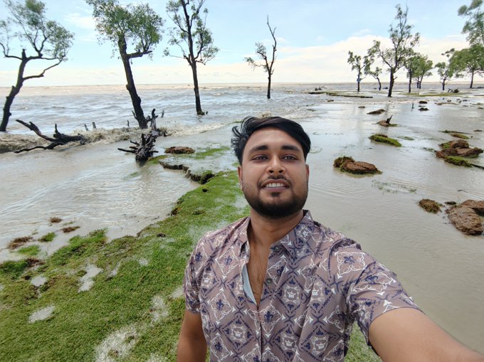 A smiling man takes a selfie in a coastal wetland area with scattered trees standing in shallow water and mossy ground. The image's outdoor, exploratory setting visually reinforces the post's theme of "exploring real life," while he promotes the @trylimitless onchain prediction market platform with a referral link.
