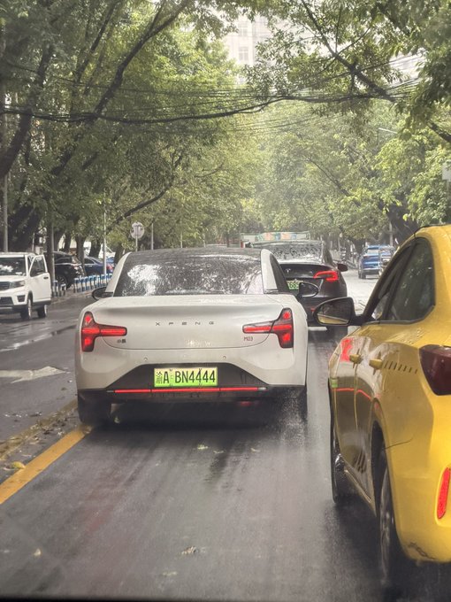 A white Xpeng P03+ electric vehicle with a green license plate reading '渝A BN4444' is stopped in traffic on a wet, tree-lined street, alongside a yellow car.