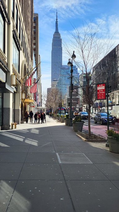 A street-level view in New York City shows the Empire State Building towering over a sunny sidewalk lined with pedestrians, street lamps, and an American flag.