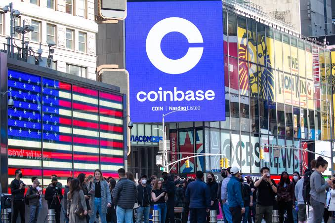 A large digital billboard in Times Square displays the Coinbase (COIN) logo with "Nasdaq Listed" beneath it, prominently featuring its recognizable "C" emblem against a bright blue background. The scene captures a bustling street with pedestrians, an electronic American flag display showing COVID-19 statistics, and a Lion King billboard, visually reinforcing Coinbase's public market presence as it launches IPO access for U.S. retail investors starting with Oura.