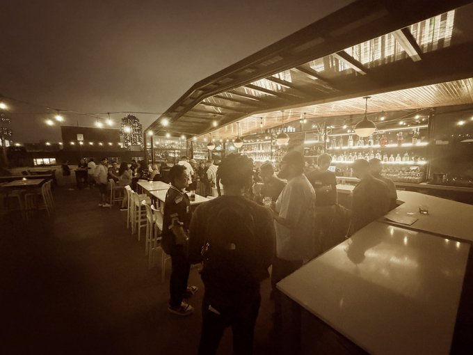 A sepia-toned photo shows a crowded rooftop bar at night, with patrons gathered around the illuminated counter under string lights.