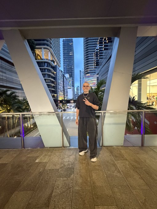A person in black clothing and sunglasses posing with a peace sign in front of an architectural structure framing modern high-rise buildings.
