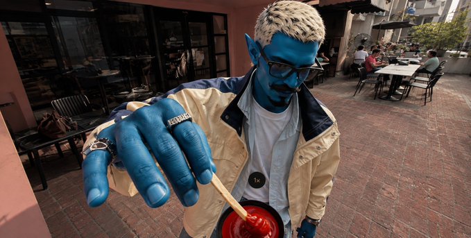 A person dressed as a blue Na'vi character from Avatar, wearing a yellow jacket and glasses, poses with an outstretched hand holding a red object.