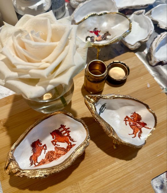 Decorative oyster shells with gold-painted edges feature painted horses and riders, displayed on a wooden surface alongside a white rose and small jars.