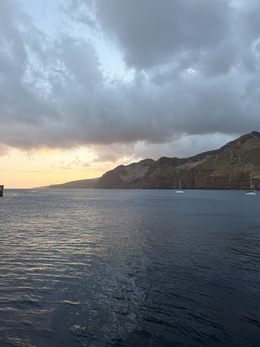 A scenic ocean view at sunset with dramatic clouds over coastal cliffs and small sailboats in the water.