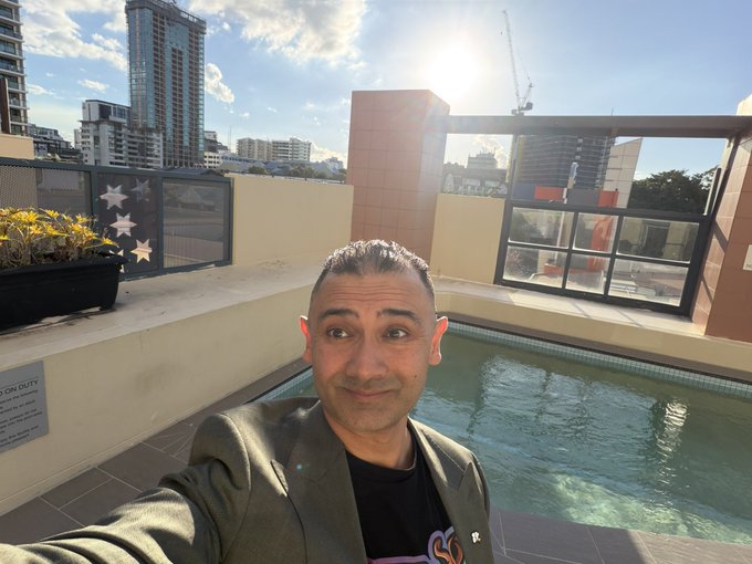 A selfie of a man in a black t-shirt and grey jacket standing next to a rooftop pool, with city buildings and a construction crane in the background.