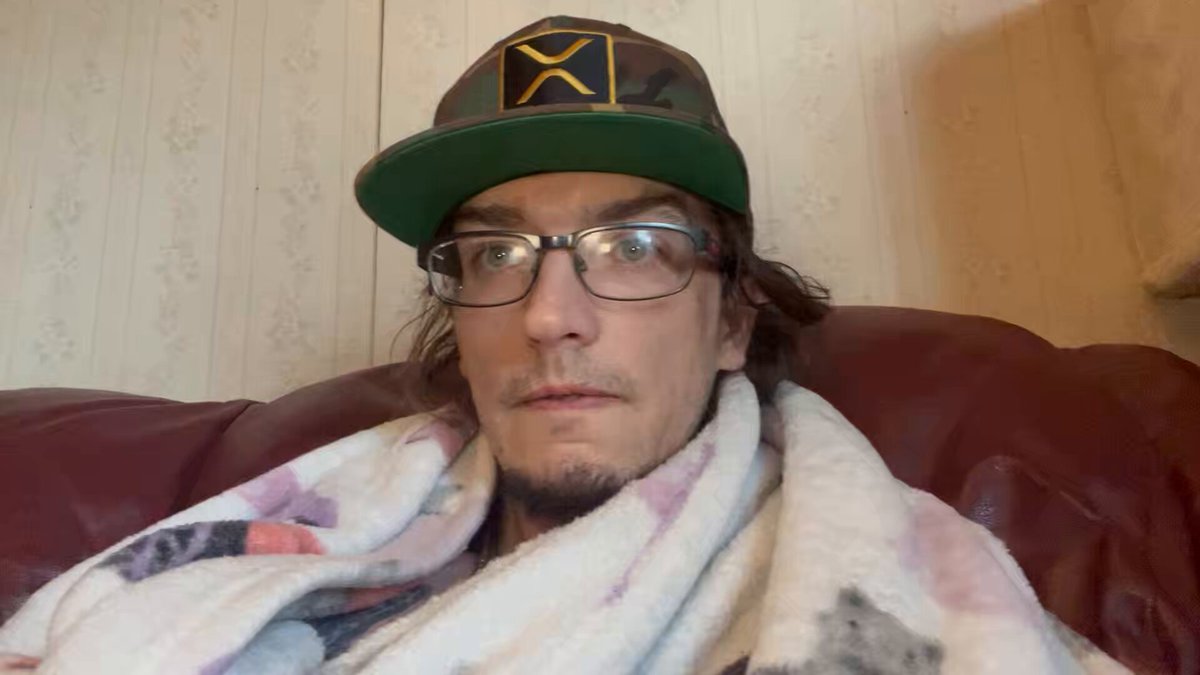 The image shows a man wearing glasses, sitting on a couch with his head resting on a blanket or towel. The background appears to be a wall, possibly indicating that he is indoors. There are no texts or other objects visible in the image, so we can only focus on the main subject - the man and his position on the couch.