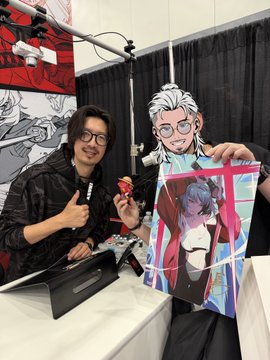 A man wearing glasses poses at what appears to be a convention or signing event, holding a small figure alongside a colorful anime-style poster.