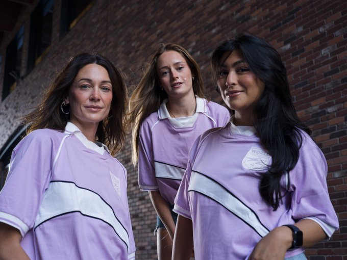 Three young women pose confidently together in matching lavender and white soccer jerseys, with a logo reading "WOMEN 42" visible on the sleeve, set against a red brick wall. The image ties to the post's call-to-action, visually reinforcing the message by featuring models representing the community waiting for the viewer to start trading on 42.