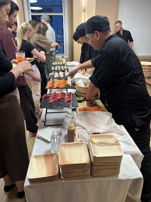 Chefs in black uniforms prepare sushi and sashimi at a long table while guests watch and serve themselves at what appears to be a catered event.