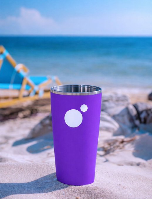 A purple tumbler cup with a white logo sits on a sandy beach with the ocean and a blue beach chair visible in the background.