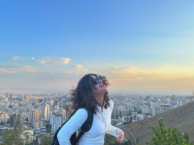 A woman with curly hair wearing sunglasses and a white jacket stands on a hilltop overlooking a sprawling city at golden hour.