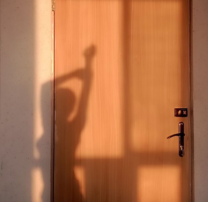 A warm-toned photograph of a shadow cast on a wooden door, showing the silhouette of a person posing with an arm raised, bathed in golden sunset light.