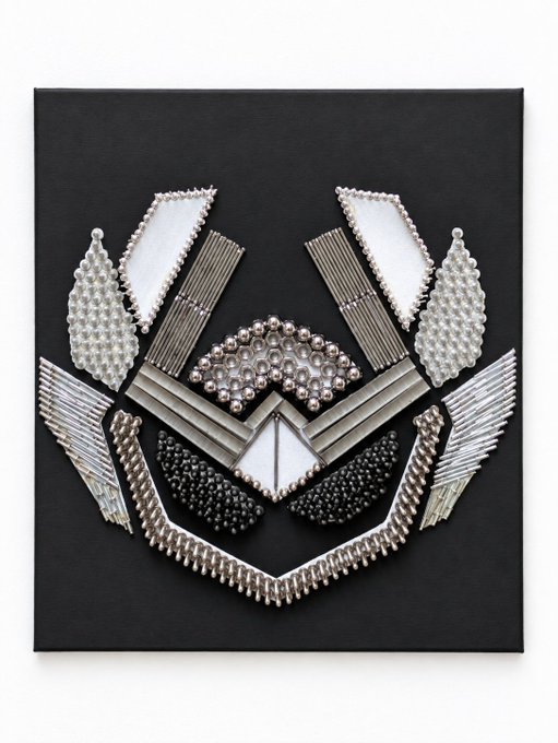 A decorative wall art piece on a black canvas, featuring a symmetrical arrangement of pearls and metallic embellishments forming an abstract crest or face-like design.