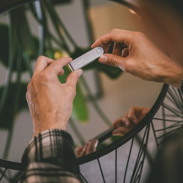The image shows a person holding up an electronic device, possibly a Ledger or similar device, with their hands. The focus of the image is on the person's hands and the device they are holding. There is no visible text related to cryptocurrency in this particular image.