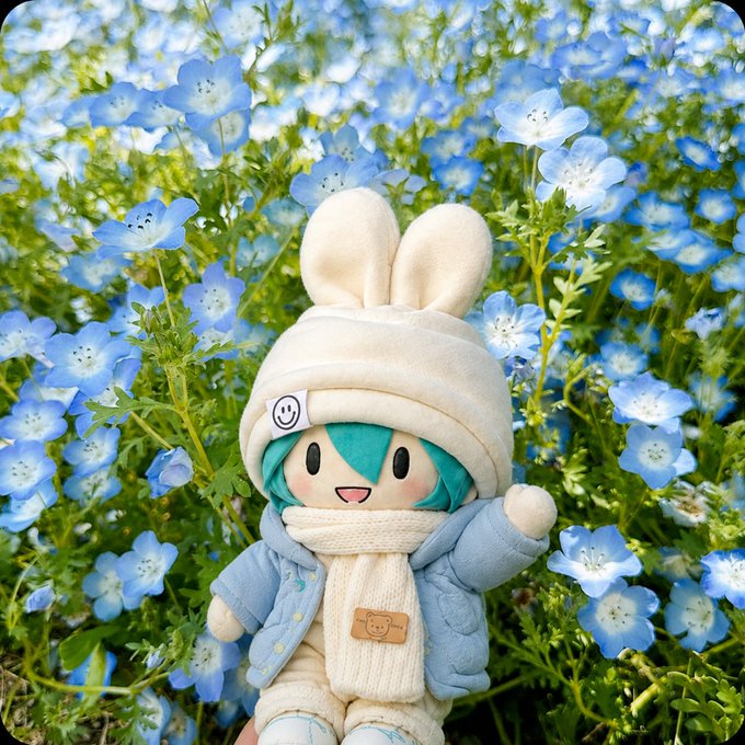 A plush doll with teal hair and a white bunny-eared hat stands smiling amid a vibrant field of blue nemophila flowers, with a small smiley-face tag visible on its hat—evoking a calm, hopeful "blue sky" morning mood that mirrors the post's sentiment of crypto markets settling into a steadier rhythm after recent turbulence.