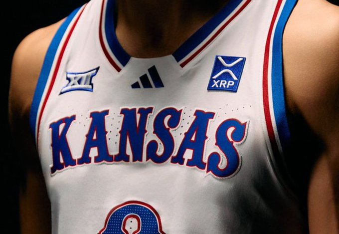 A close-up of a white Kansas Jayhawks basketball jersey with blue and red trim, featuring the Adidas logo, Big 12 conference logo, and an XRP cryptocurrency patch on the chest.