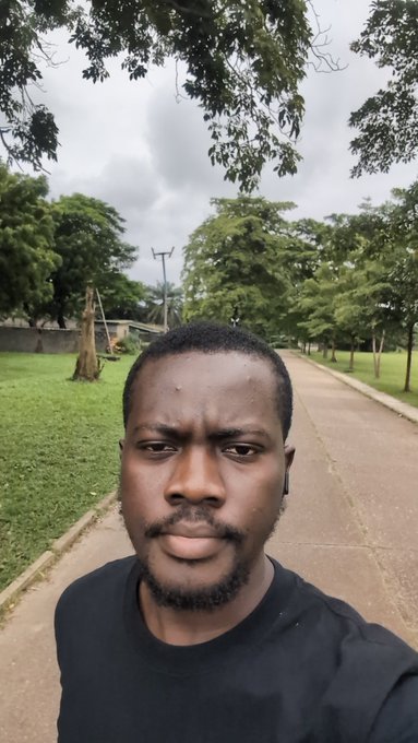 A selfie of a bearded man in a black t-shirt standing on a paved pathway lined with green trees and grass under an overcast sky.