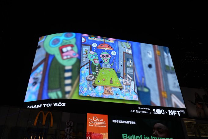 A nighttime photo of a billboard displaying NFT artwork by J.P. MoreGain via 100xNFT, showing cartoon zombie-like characters, located above a McDonald's and Cane Sweet cafe.