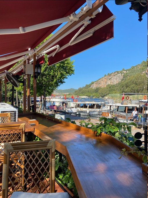 A view from a waterside cafe or restaurant showing wooden tables and chairs under a red awning, looking out at tour boats docked near a rocky coastline.