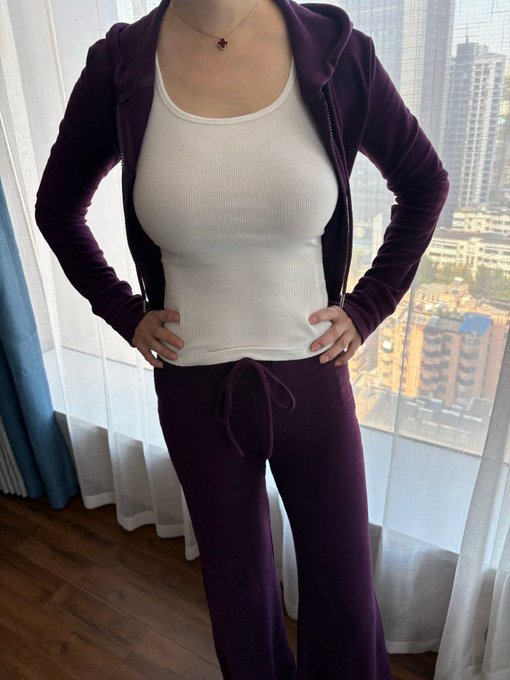 A person poses in a matching deep purple velour zip-up hoodie and drawstring pants set over a white ribbed tank top, standing near a window overlooking a city skyline. The image reflects the celebratory tone of returning to the platform after a freeze, showcasing a stylish "long time no see" outfit reveal.