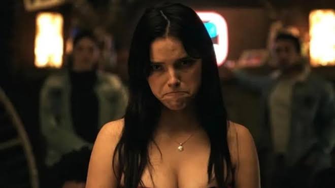 A young woman with long dark hair and a pearl necklace glares intensely at the camera in a dimly lit room with two men blurred in the background.
