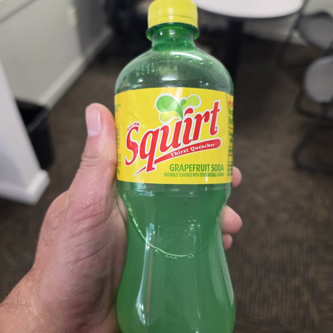 A hand holds up a green plastic bottle of 'Squirt Thirst Quencher Grapefruit Soda' in an indoor setting with chairs visible in the background.