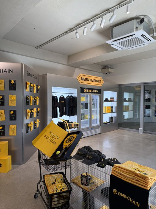 The photo shows the interior of a "MERCH MARKET" branded BNB Chain store, featuring yellow and black merchandise including t-shirts, hats, and tote bags, with a shopping cart displaying a "SHAPE THE FUTURE" tote that reinforces the post's promotional theme for BNB Chain.