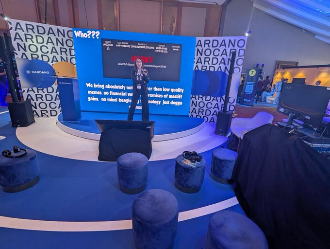 A woman presents at a Cardano event on a stage decorated with blue carpet and ottomans, with screens displaying 'CARDANO' and 'Who???' branding.