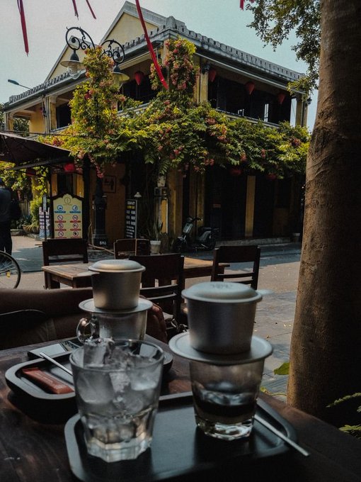 A wooden table holds two traditional Vietnamese drip coffee filters over glasses of ice, set against the backdrop of a yellow building draped in greenery with red lanterns hanging above.
