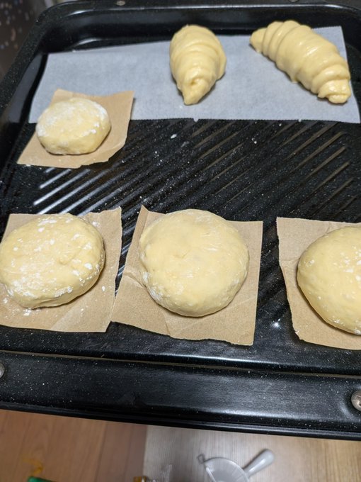 Several pieces of raw dough, including round buns and croissants dusted with flour, rest on parchment paper squares on a black baking tray.