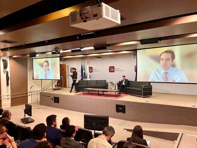 A panel discussion takes place at DCGG Brazil, where Sam Elfa, Community Spokesperson of the TRON DAO, speaks on stage with his image projected on large screens behind him, addressing an audience about blockchain transparency, stablecoins, and the TRON ecosystem. The backdrop displays the USP (Universidade de São Paulo) logo, reflecting the event's academic setting for this talk on Web3 adoption.