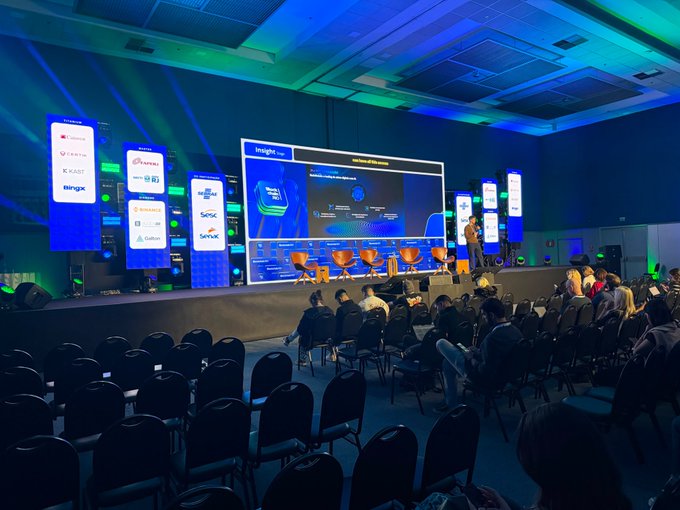 A conference room is filled with an audience watching a keynote presentation on a large screen, with vertical banners displaying logos like Binance and OKX.