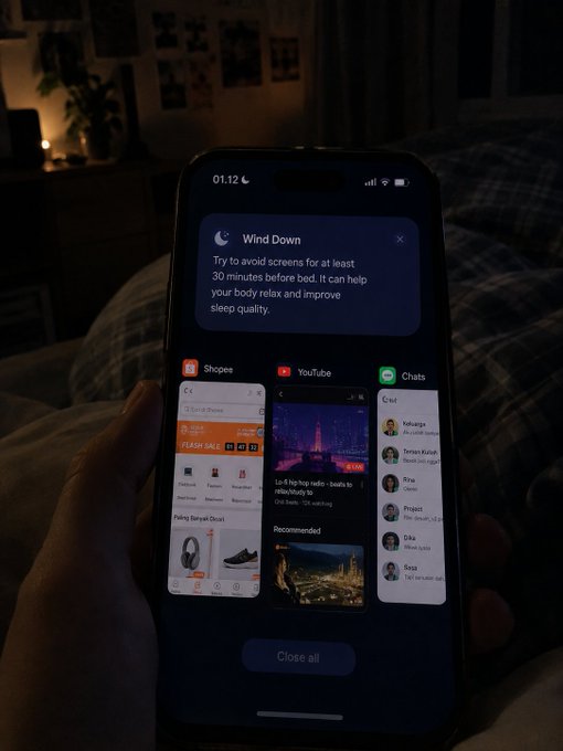 A hand holding a smartphone at 01:12 displaying a "Wind Down" notification about avoiding screens before bed, with the iOS app switcher open showing Shopee, YouTube, and Chats.