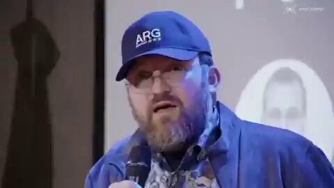 The image shows a man wearing glasses and a blue baseball cap with the word "ARG" written on it. He appears to be speaking into a microphone, possibly at an event or gathering. The background of the image features a projection screen displaying a picture of a person's face, which is likely related to the content being discussed by the speaker.