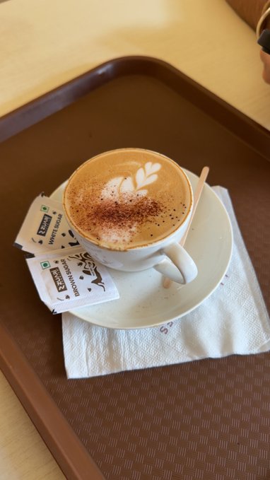 A cappuccino with leaf latte art and cocoa powder dusting sits on a saucer atop a napkin on a brown tray, accompanied by white sugar packets labeled 'Green Card White Sugar'.