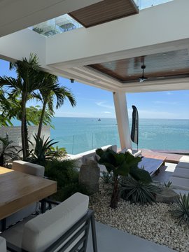 View from a modern outdoor patio overlooking a beach and ocean, featuring tropical plants, lounge chairs, and a wooden dining table.