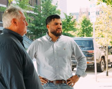 Two men stand outdoors having a conversation on a sunny day, with a parked vehicle visible in the background.