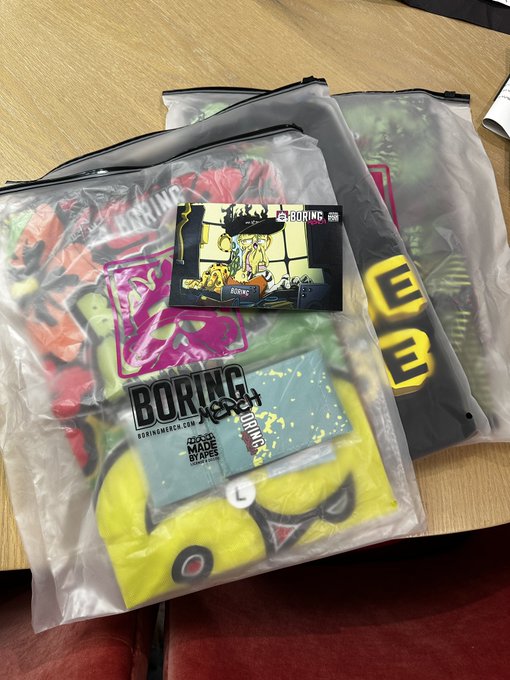 Clear plastic bags containing streetwear clothing with 'BORING' branded tags and graphics, including a yellow smiley face design, resting on a wooden surface.