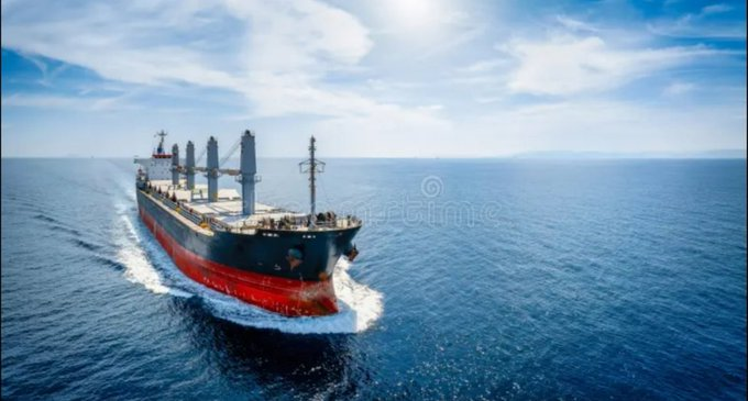 An aerial view of a red and white cargo ship sailing through blue ocean waters under a sunny sky with scattered clouds.
