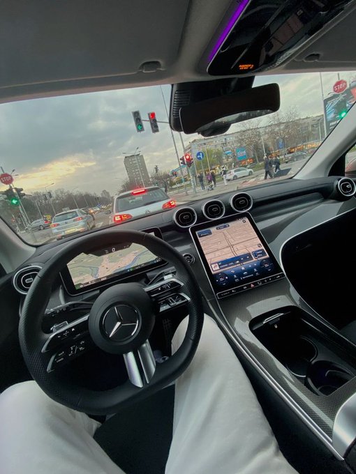 Interior view from the driver's seat of a Mercedes-Benz car showing the steering wheel, dashboard screens with maps, and a street view with traffic lights and cars ahead.