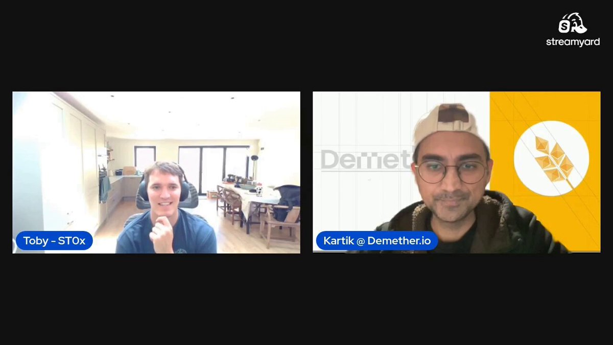 The image shows two men sitting at separate tables, both wearing headphones and looking at their computer screens during an online meeting or video call. The man on the left has his hand raised, possibly asking a question or making a point. The other man is smiling, indicating that he is engaged in the conversation. The background of the image reveals a kitchen area with a dining table and chairs, suggesting that they are likely having their meeting over dinner or while enjoying some food together before continuing their discussion.